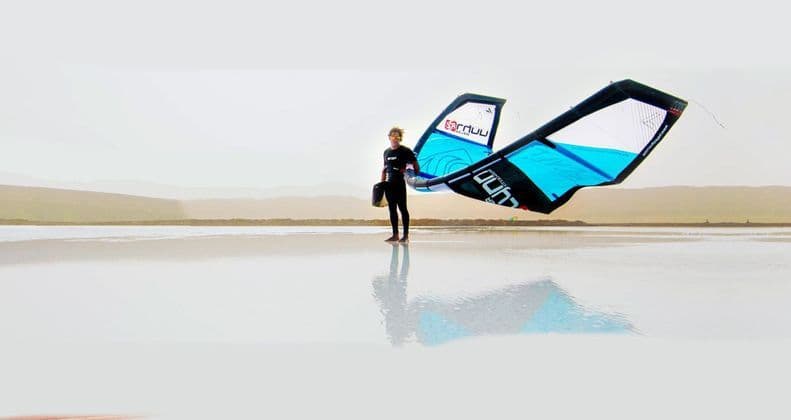 Una persona con una muta da sub tiene un grande aquilone da kitesurf blu e nero, stando in piedi in acqua bassa e riflettente.
