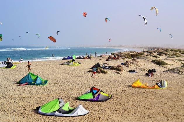 Una vasta spiaggia sabbiosa è animata da persone che fanno kitesurf, con molti aquiloni colorati sulla sabbia e nel cielo sopra l'oceano.