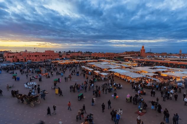 Ein belebter Stadtplatz in der Abenddämmerung, von oben gesehen, voller Menschen und beleuchteter Marktstände.