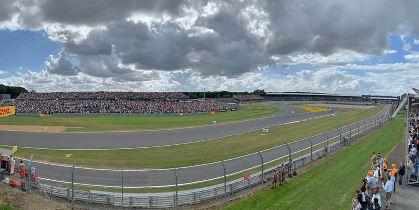 A panoramic view of a racetrack circuit with large crowds filling the grandstands under a cloudy sky.