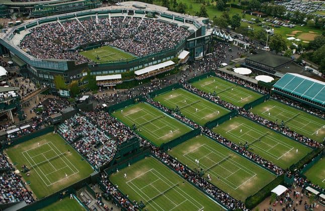 Aerial view of a packed tennis stadium and numerous grass courts with ongoing matches and crowds of spectators.