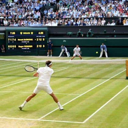 Roger Federer hits a forehand against Novak Djokovic during a tennis match on a grass court in front of a crowd and scoreboard.