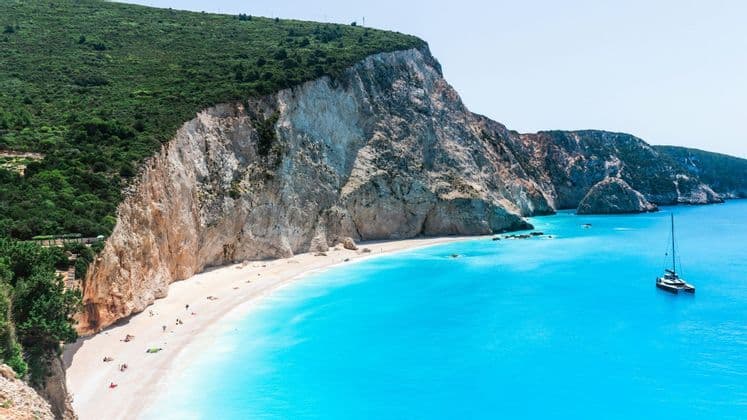 Veduta aerea di una spiaggia isolata di sabbia bianca e acqua turchese, ai piedi di una grande scogliera dalla cima verde, con un catamarano che naviga nelle vicinanze.