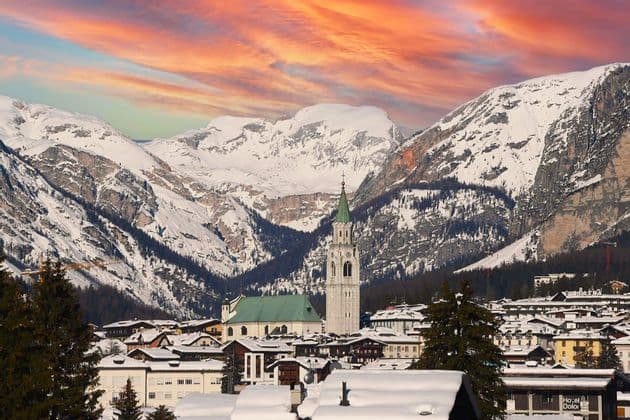 Un villaggio innevato con una chiesa imponente è adagiato tra grandi montagne innevate sotto un cielo colorato al tramonto.