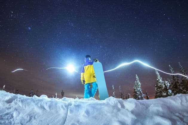 Uno snowboarder sta su una collina innevata di notte, tenendo uno snowboard sotto un cielo pieno di stelle e scie luminose.