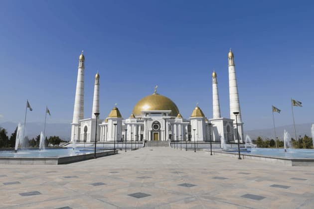 Una grande moschea bianca con una cupola dorata e quattro minareti si trova dietro una piazza pavimentata con fontane sotto un cielo azzurro e limpido.