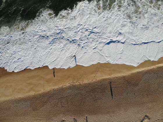 An aerial view of a sandy beach with 'WEROAD' written in the sand, while people stand near the shoreline as a wave crashes.
