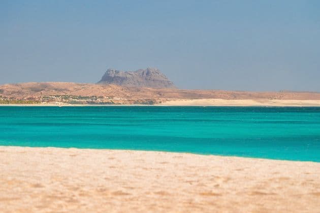 Une vue paysagère depuis une plage de sable, regardant par-dessus une eau turquoise vers une côte aride lointaine avec une montagne rocheuse.