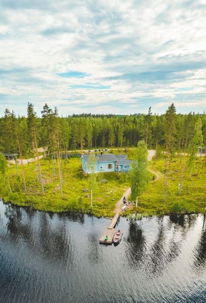 Eine Luftaufnahme eines blauen Holzhauses an einem grasbewachsenen Seeufer, mit einem Steg, zwei kleinen Booten und einem umliegenden Kiefernwald.