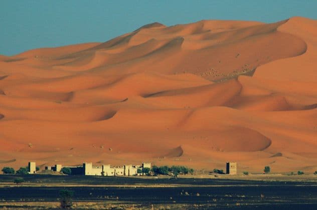 Un piccolo insediamento di edifici in adobe e alberi si trova alla base di imponenti dune di sabbia arancione sotto un cielo azzurro e limpido.