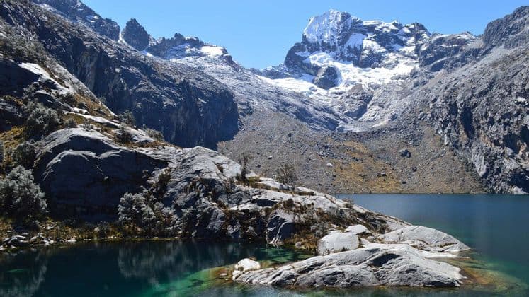 Un limpido lago alpino turchese con sponde rocciose giace ai piedi di imponenti montagne innevate sotto un cielo azzurro.
