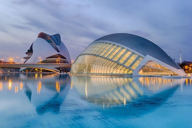 Dos edificios arquitectónicos modernos e iluminados reflejados en un estanque de agua tranquila al anochecer.