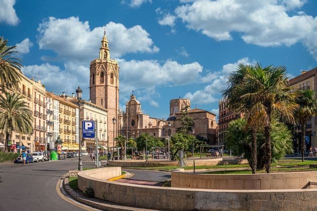 Una catedral de piedra con un campanario alto y ornamentado domina una soleada plaza de la ciudad con una rotonda y palmeras.