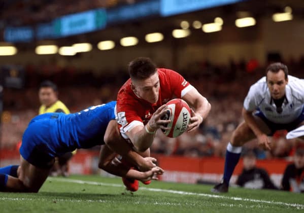 Un giocatore di rugby in maglia rossa si tuffa per segnare una meta, mentre viene placcato da un avversario in maglia blu.