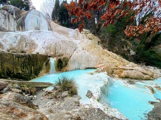 Terrazze di travertino bianco con piccole cascate che sfociano in piscine di acqua termale turchese, incorniciate da alberi con foglie autunnali.