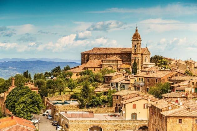 Un borgo in pietra con tetti in terracotta e un campanile di chiesa sorge su un verde pendio, con montagne in lontananza.
