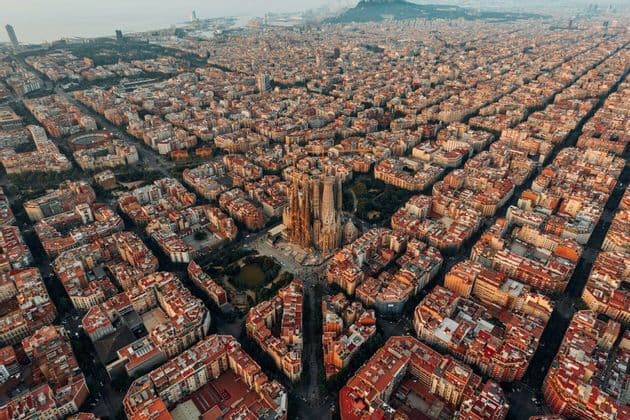 Vue aérienne de la cathédrale de la Sagrada Família entourée par les rues quadrillées et les bâtiments d'une ville tentaculaire au coucher du soleil.