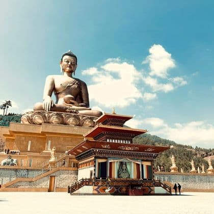 Eine massive bronzene Buddha-Statue sitzt auf einem Thron und blickt auf einen kleineren, verzierten Tempel in einem großen Innenhof.