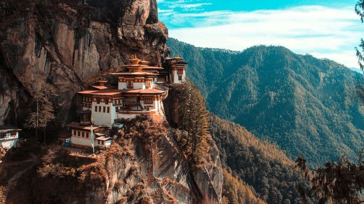 Ein Klosterkomplex mit weißen Mauern und roten Dächern schmiegt sich an eine steile Klippe und überblickt bewaldete Berge unter blauem Himmel.