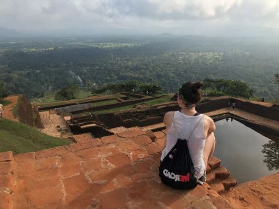 Una mujer con una mochila WeRoad se sienta en una estructura de piedra elevada, con vistas a un vasto valle verde y ruinas antiguas.