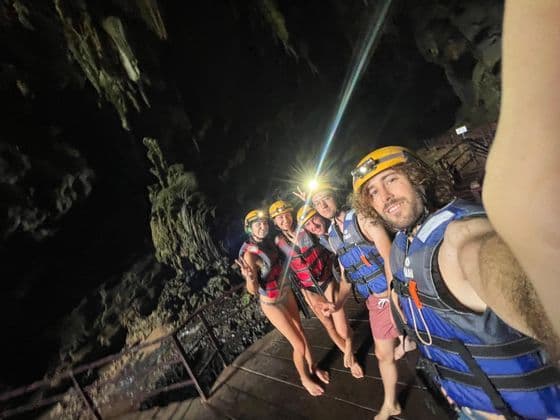 Un viaggio di gruppo WeRoad con caschi, lampade frontali e giubbotti di salvataggio posa per un selfie su una passerella di legno all'interno di una grotta buia.