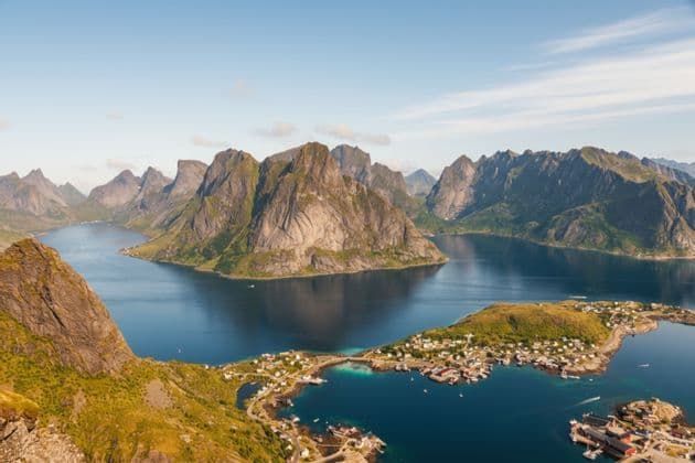Una vista aerea di un villaggio costiero ai piedi di grandi montagne rocciose circondato da un fiordo blu profondo.
