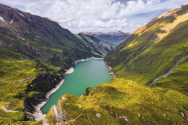 Vista aerea di un lago alpino turchese incastonato in una profonda valle tra ripide montagne verdi, sotto un cielo nuvoloso.