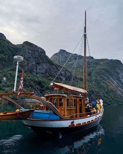 Eine WeRoad Gruppenreise auf einem Holzsegelboot fährt entlang eines Fjords mit steilen, felsigen Bergen im Hintergrund.