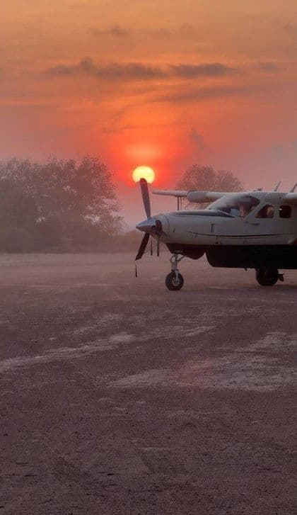Un piccolo aereo ad elica parcheggiato su un campo polveroso al tramonto, con il sole arancione che splende dietro la sua elica.