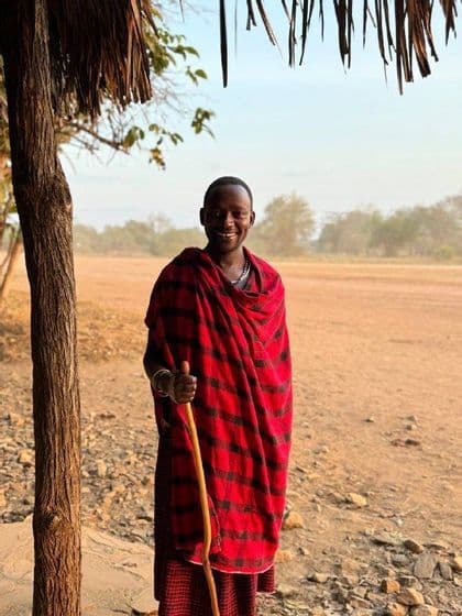 Un uomo sorridente con una tunica scozzese rossa e nera tiene un bastone da passeggio mentre è in piedi accanto a un albero in un paesaggio polveroso.
