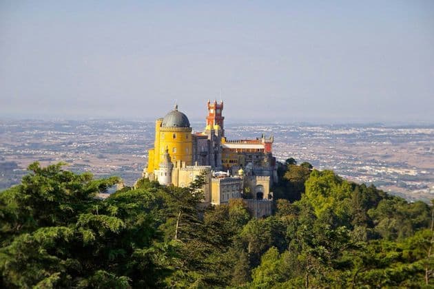 Un castello variopinto con torri gialle e rosse si erge su una collina boscosa, dominando un vasto paesaggio.