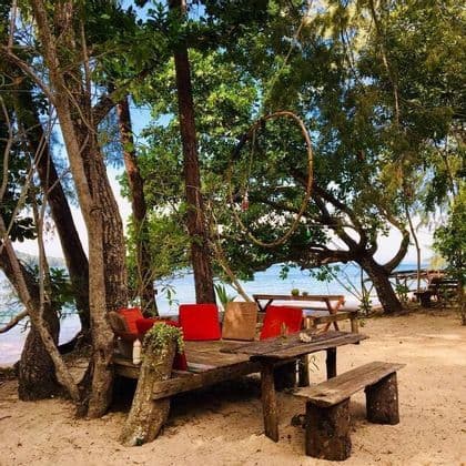 Un coin salon rustique en bois avec des coussins colorés sur une plage de sable, au milieu d'arbres verdoyants surplombant la mer.