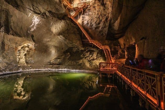A large, illuminated cavern with a wooden staircase climbing the rock wall and a walkway next to a dark pool of water.