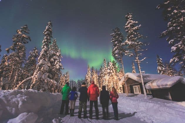 Eine WeRoad-Gruppenreise steht nachts in einem verschneiten Wald und beobachtet die Nordlichter über den schneebedeckten Bäumen und einer Holzhütte.