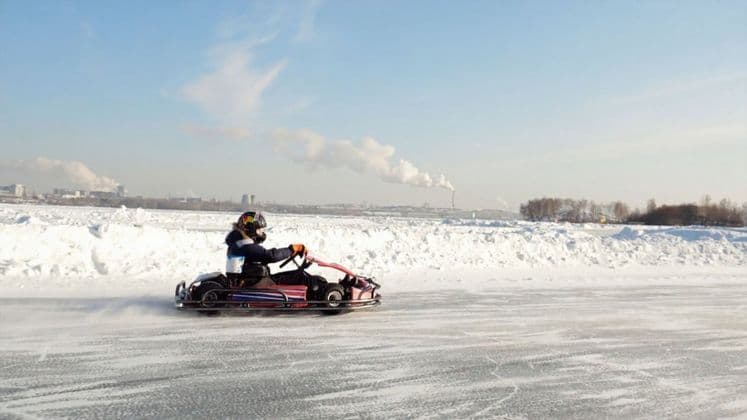 Eine Person mit Helm fährt Go-Kart auf einer gefrorenen, schneebedeckten Strecke, vor einer fernen Stadtsilhouette unter blauem Himmel.