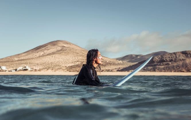 Una persona con i capelli bagnati in una muta nera sorride mentre è seduta su una tavola da surf nell'oceano, con colline sabbiose sullo sfondo.