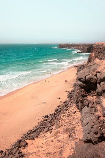 Una vista da una scogliera rocciosa che si affaccia su un'ampia spiaggia sabbiosa con acqua turchese e piccole onde che si frangono sulla riva.