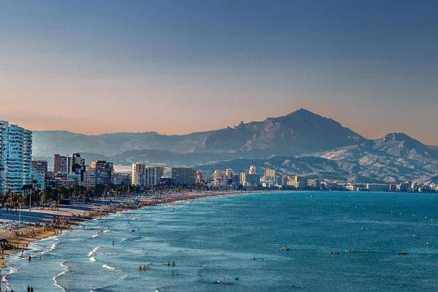 Un paesaggio urbano su una costa con una spiaggia affollata dove le persone nuotano nell'oceano, con grandi montagne visibili in lontananza.