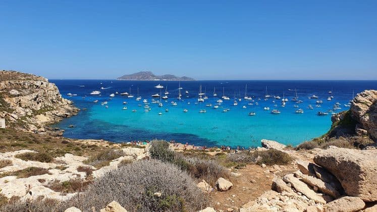 Vista dall'alto di una costa rocciosa con baia turchese e numerose barche ancorate, e un'isola in lontananza.