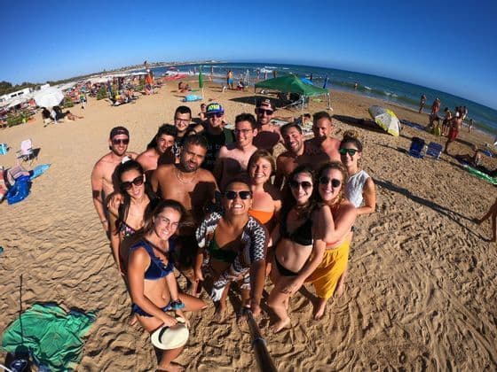 Un groupe WeRoad en voyage prenant un selfie grand-angle sur une plage ensoleillée et bondée, au bord de la mer.