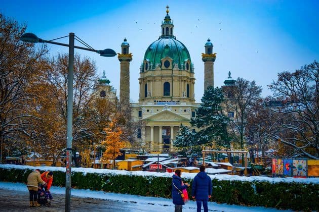 Eine große Kirche mit grüner Kuppel überragt einen verschneiten Park, in dem Menschen über einen Wintermarkt schlendern.