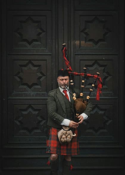Ein Mann in traditioneller schottischer Tracht, mit Kilt und Tweedjacke, hält einen Dudelsack und steht vor einer großen, verzierten Holztür.