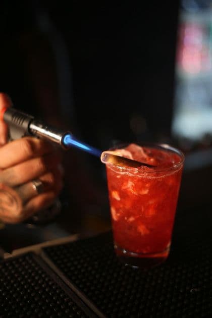 Un barman con una mano tatuada usa un soplete de cocina para encender una rama de canela en un cóctel rojo con hielo picado.