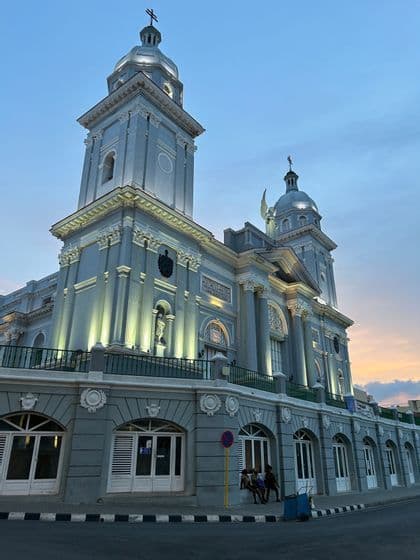 Una ornamentada catedral gris con dos torres está iluminada por focos bajo un cielo crepuscular.
