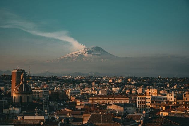 Un paesaggio urbano con un edificio a cupola si trova sotto un grande vulcano innevato che emette una colonna di fumo contro un cielo color verde acqua.