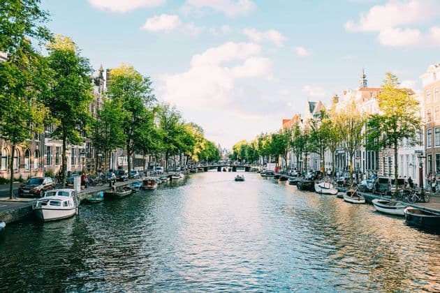 A wide city canal flows between streets lined with traditional buildings and green trees. Numerous boats are moored along the banks on a sunny day.