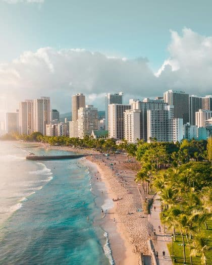 Una vista aerea di una spiaggia cittadina affollata con acqua turchese e palme, sullo sfondo di moderni grattacieli.