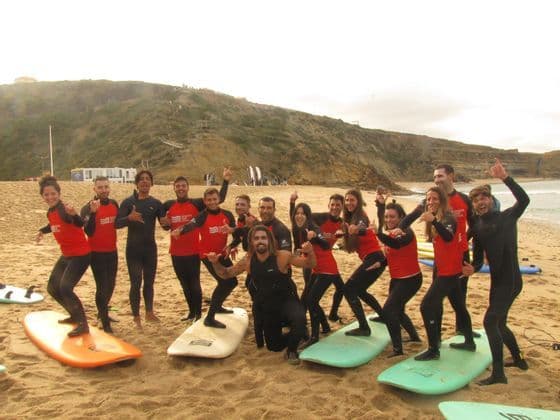 Un gruppo WeRoad in muta posa per una foto con tavole da surf su una spiaggia sabbiosa di fronte a una scogliera.