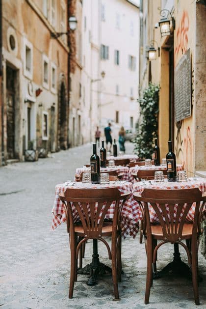 Tables at an outdoor restaurant with red checkered cloths and wine bottles are set on a narrow cobblestone street.
