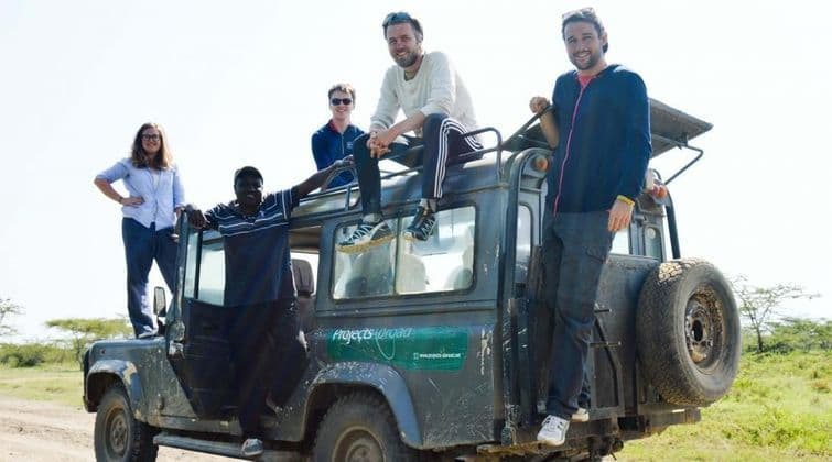 Un groupe WeRoad de cinq personnes souriant et posant sur une jeep safari verte en savane.